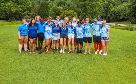 Group of diverse people with arms around one another on green field smiling for the camera.