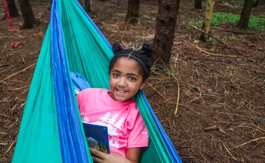 Girl laying in green hammock with book open in hand and smiling for the camera