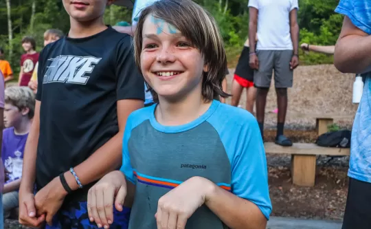 Boy doing funny pose with hands and with a genuine big smile, he's also wearing face paint and you can see several other children in the background.