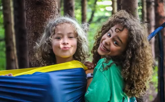 Forested background, hammock between two trees, two girls exuberantly smiling at the camera, one in the hammock