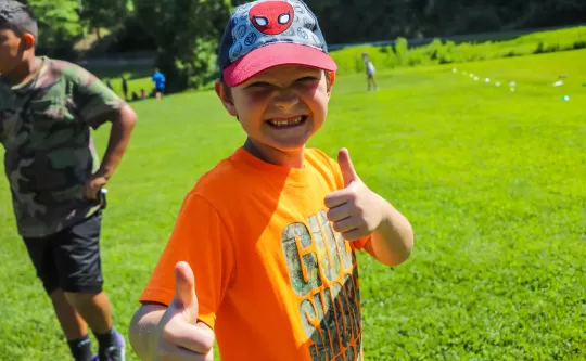 Boy with a baseball hat and large smile on his face standing in a field giving a thumbs up. There is another kid in the background walking off screen.