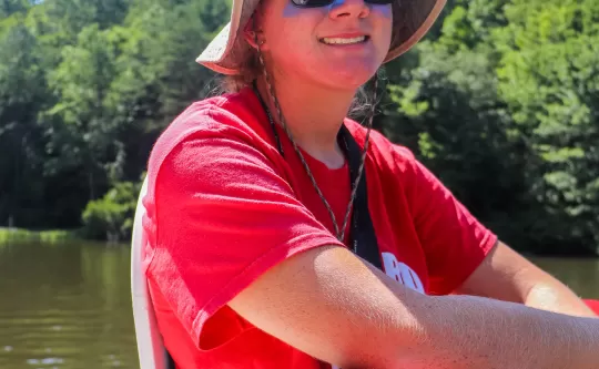 Lifeguard on stand with flotation device held in hand, hat on head, smile on face, lake in background.