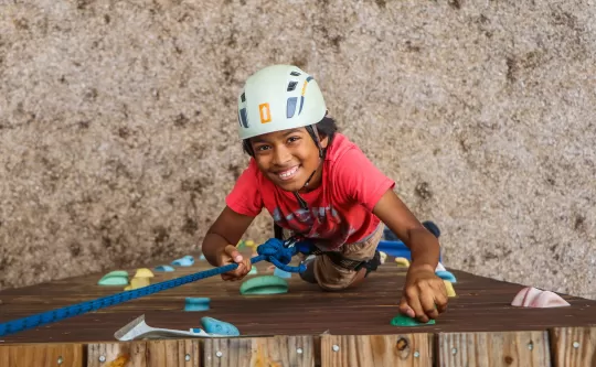 Child climbing a rock climbing wall looks at camera with a big smile on face. 